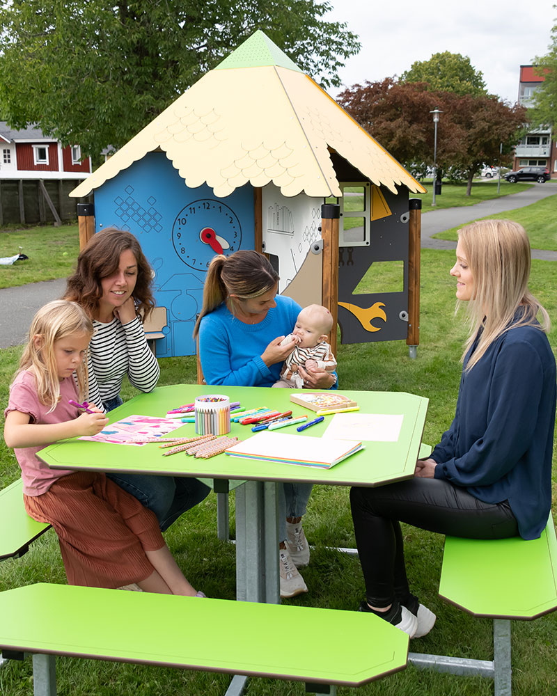 Un groupe de femmes avec des enfants se détend sur une table de pique-nique HPL verte, qui donne sur un skate park où jouent des enfants.